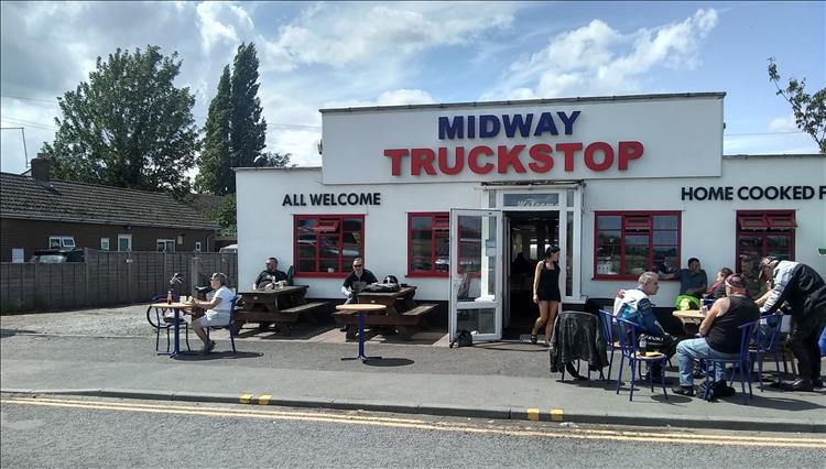 A white building with big signage declaring Midway Truck Stop. The sun is shining and a few people sit outside