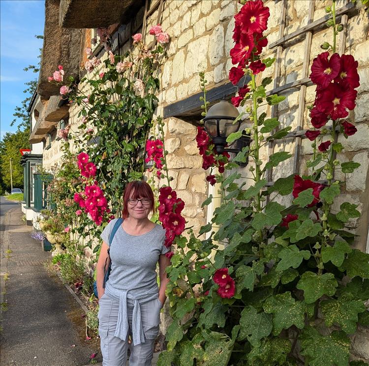 Sharon next to a thatched cottage with vibrant red flowers growing on the wall