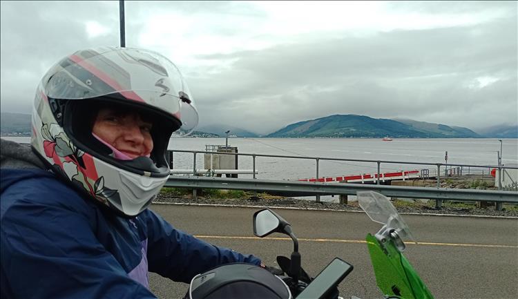 Sharon's on her bike with her helmet on smiling at the camera at the ferry terminal with dramatic scenery behind her