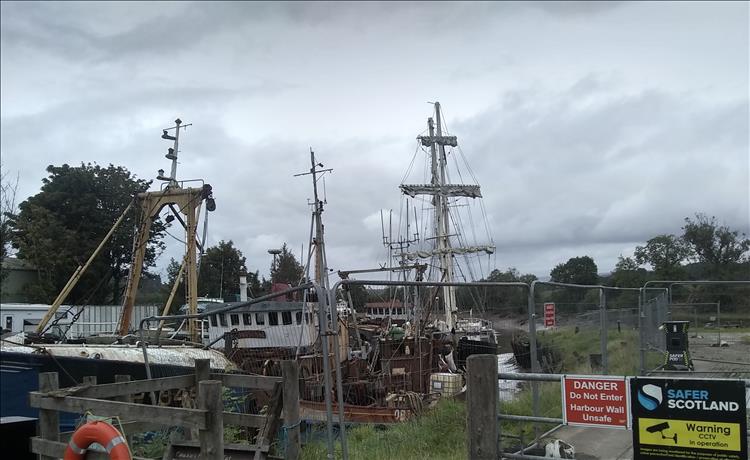 Behind the rusty trawlers, wire fencing, and run down wall we see the masts of a sailing ship