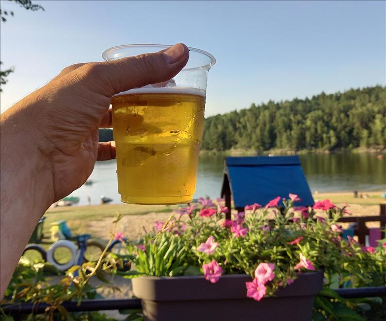 A hand holds up a cold beer, behind flowers and the lake and sunshine