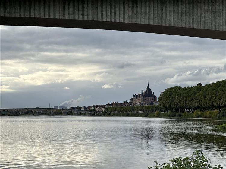 From under the concrete arch of a bridge is a broad river and a castle with spires across the water