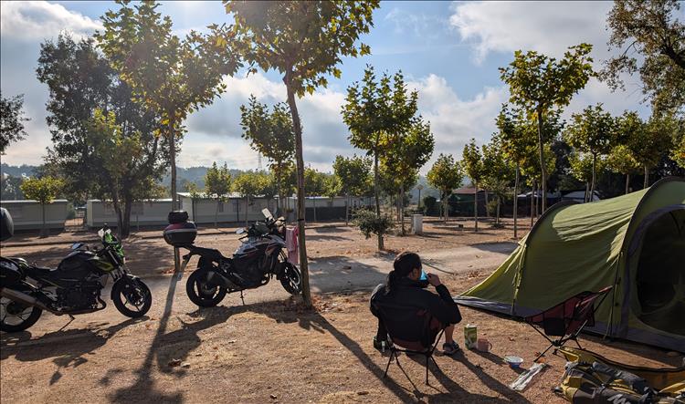 Ren sits in a chair drinking, the campsite is deserted and the sun is shining