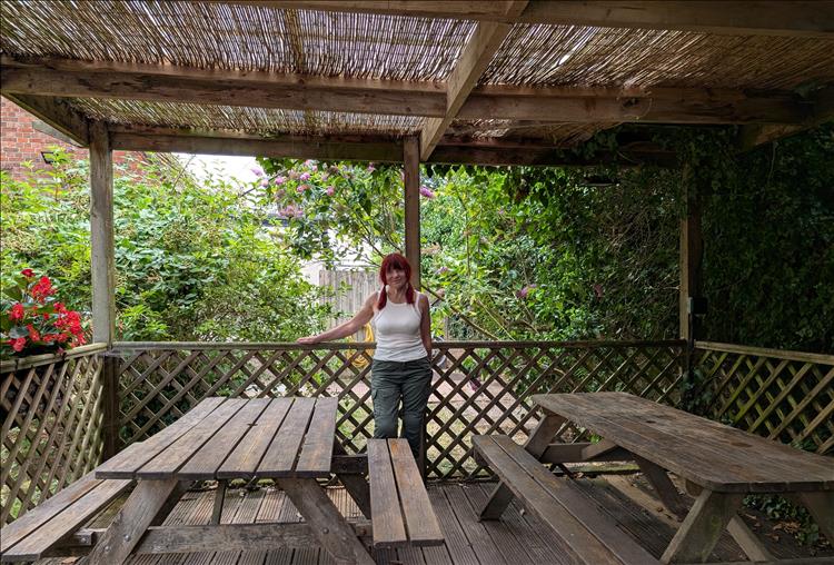 Sharon beside wood bench tables, under a rattan shelter waiting for her brew