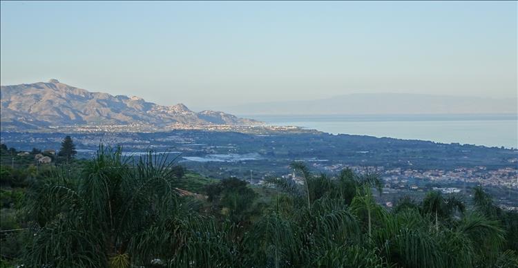 Looking from a mountain across flat valley with towns to another mountain, the sea and in the far distance more land