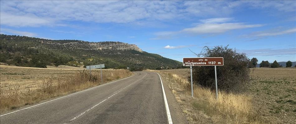 sign reads Hortezuelos 1137m beside an empty road with a tree covered rocky outcrop ahead
