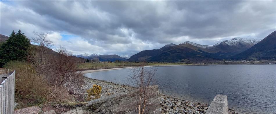 Rugged highland scenery, a calm loch, hills and snow capped mountains, heavy clouds, and hardy trees