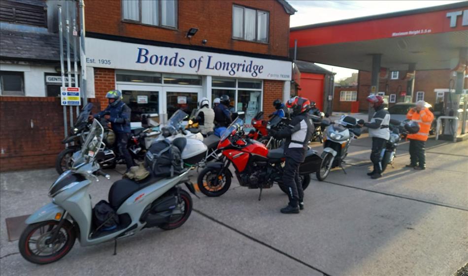 A number of riders and motorcycles putting their gear on beside a petrol station