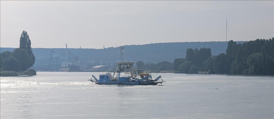 A very broad river, large boats and cranes in the hazy distance and a small blue car ferry