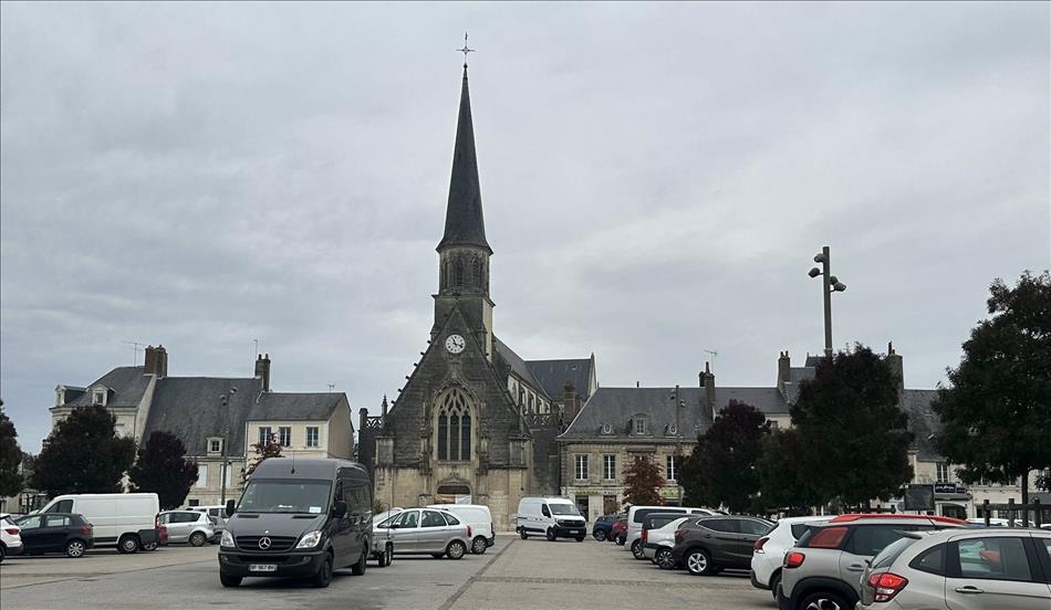 A car park witha  few vehicles, a stone church with a tall narrow spire and a few other buildings