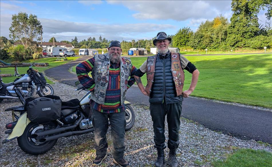 2 older chaps with cut offs covered in patches pose in a silly manner beside their cruiser motorcycles