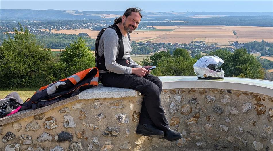 Ren sits on wall wearing black heavy cotton trousers and squinting in the sun