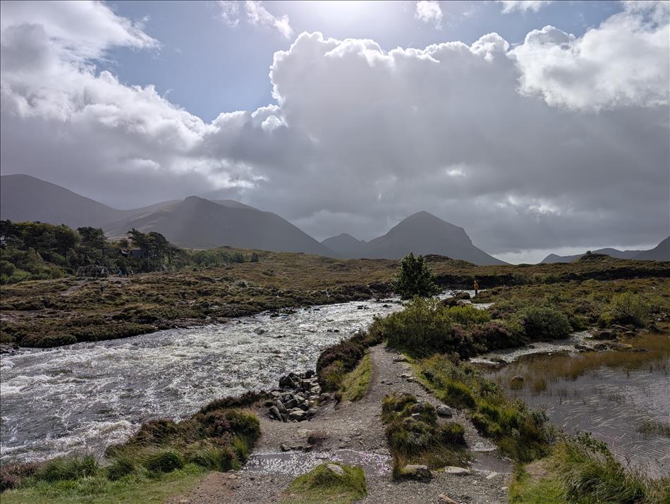 A bubling river runs through a scene with angular mountains and heavy clouds although the sun shines through